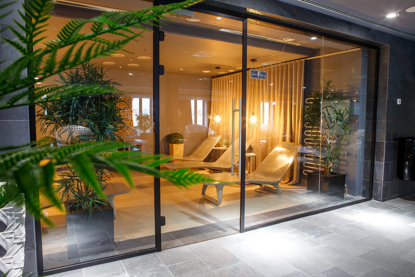 Modern spa relaxation room with grey lounge chairs and green plants.